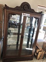 Front view of wooden vintage display cabinet showing glass doors, glass shelves inside and mirrored back panel reflecting room light. Carved wood crest on top is detached but shown separately in a close-up photo.