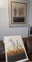 Photo showing one framed print hanging on the wall featuring tall leafless trees and another leaning against a brown couch.