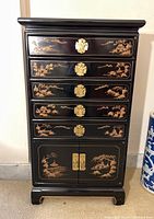 Front view image showing the black lacquer jewelry chest with detailed gold chinoiserie landscape decoration, five drawers with brass lock plates, and bottom two-door cabinet with brass handles.