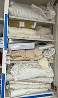 Shelves containing folded and rolled bed linens and towels in mainly white, cream, and beige colors, stacked in an enclosed shelving unit.