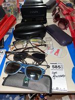 Multiple pairs of sunglasses and reading glasses arranged with cases including a Prada branded case and various frames in black, blue, and tortoise colors