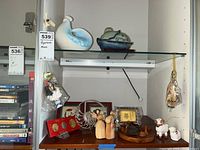 Two shelves with various figurines including ceramic pieces, corn husk dolls, wooden tray with carvings, coin set, and ceramic sheep figurine