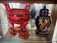 Red cinnabar tripod incense burner missing lid on wooden carved stand beside a blue ginger jar with peacock design on dark wooden stand