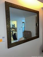 Full view of the rectangular mirror mounted on a white wall, showing the bronze mosaic frame and beveled mirror reflecting a kitchen and hallway.