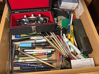 Office supplies items including magnifying glasses in a case, assorted pens and markers, stapler, ink cartridge, and a trackball mouse all in a drawer.