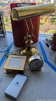 Photo showing brass adjustable desk lamp, silver quartz clock, small gold-tone picture frame, and red clipboard with red metal waste can in background.