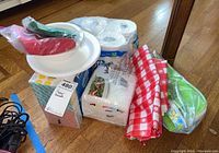 Paper goods including multi-pack toilet tissue, box of facial tissues, stack of white disposable plates, red and blue plastic utensils, red and white checkered picnic cloth, and insulated floral picnic cooler bag.