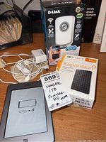 Overview photo showing all items together on a desk: Amazon Kindle e-reader, D-Link wireless camera in box, Seagate 1TB portable hard drive in box, and cables including a LapLink transfer cable.