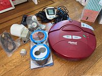 Red iRobot Roomba vacuum with spot and clean buttons alongside blood pressure monitors and accessories on wooden floor.