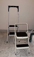 Tall two-step ladder with black handle and black steps alongside shorter white frame two-step ladder with black steps and small handle bar, on marble floor.