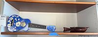 Side view of blue ukulele with white floral designs and wooden fish next to it