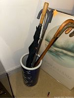 Full view of umbrella stand with black umbrella, multiple canes inside and part of nearby framed art.