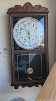 Front view of vintage wooden framed wall clock showing ornate carved top, glass panels, clock face with 12-hour and 31-day calendar markings, and brass pendulum.