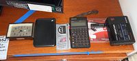 Photo showing alarm clock, black case, Sony microcassette recorder, HP calculator, boxed USB flash drive, and Sony Walkman on wooden surface.