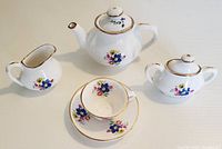 Full set showing teapot, sugar bowl, creamer, cup and saucer with floral pattern and gold trim on white ceramic.