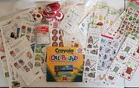 Photo showing an assortment of craft stickers sheets, rolls of red and white striped tape, packet of MACO tags with string, and Crayola oil pastels box.