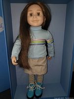 Doll standing inside original blue Maplelea box wearing khaki skirt, light blue striped turtleneck and blue hiking shoes