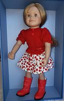 Front view of 18 inch Maplelea Leonie doll with blonde hair, red sweater, cherry skirt, and red boots inside blue box.