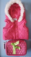 Pink sleeveless ski jacket with faux fur hood trim and pink mini polka dot suitcase with green bow, wheels and handle, laid flat.