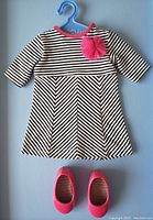 Photo of black and white striped doll dress with pink flower detail and matching pink shoes on a blue hanger