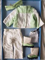 Photo of doll ski outfit with light green and white jacket, white pants, green and beige boots, head warmer, white sunglasses, and carrying pouch inside blue box.