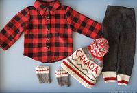 Photo showing the full set of doll clothes including the red and black plaid shirt, grey pants, Canada toque, socks, and mittens laid out flat against a blue background.