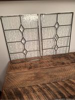 Two clear stained glass window panels with rippled and beveled geometric diamond design, standing upright on a rustic wooden surface