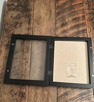 Open black wooden shadow box frame showing interior backing and small sealed bag of pearl pins attached.