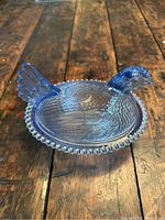 Perspective view of blue glass hen dish on wooden surface, highlighting the textured wings, tail feathers, and beaded edge of the dish.