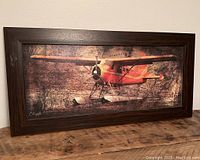 Framed print showing red float plane on textured Canada map background, placed on wooden surface.
