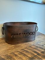 Front angled view of oval metal firewood tub with 'FIREWOOD' cutout and handles on the sides.
