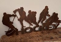 Front and angled view of copper colored metal wall decor showing five hockey players and word 'HOCKEY'. The decor is standing on a wooden surface.