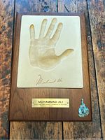 Front view of Muhammad Ali handprint plaque showing raised handprint, signature, brass nameplate, and boxing glove emblem on wood plaque.