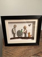 Framed pebble art showing the full composition of a couple with a baby in stroller, dog, and bird inside a black shadow box frame.