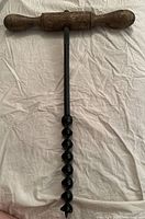 Full view of primitive wood auger hand drill with T-shaped wooden handle and spiral iron bit on fabric background.