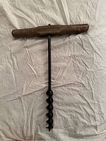Full view of the primitive wood auger hand drill showing T-shaped wooden handle and metal spiral bit on cloth background.