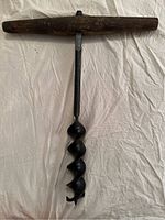 Photo showing primitive wooden T-handle attached to large black metal spiral drill bit of one hand auger drill.