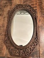 Full view photo of the oval mirror with ornate copper-color frame placed on rustic wooden surface.