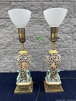 Front view of two ceramic and metal lamps with brass bases and cherub figures, showing floral openwork ceramic detail and white glass shades.