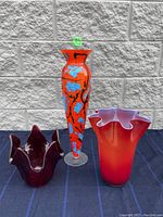 Three glass vases placed side by side against a gray stone wall background: tall orange painted vase with blue floral patterns; medium red-orange gradient ruffled vase; deep red multi-peak vase