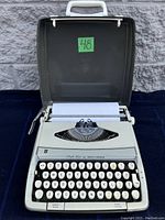 Photo showing the closed carrying case open with the typewriter inside loaded with blank paper. The typewriter's keys and platen roller are visible.
