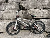 Side view of white DK Drew Bezanson BMX bike leaning against a stone wall outdoors. Shows full bike profile, frame, tires, and pedals.