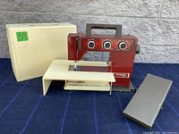 Full view of the Husqvarna 5610 sewing machine in red with cream base, cream packaging box with green sticker, and grey foot pedal on blue fabric