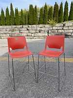 Pair of mid-century modern style bar stools with red plastic seats and backs and chrome sled legs set outside on pavement with stone wall background.