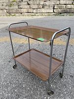 Side-angle showing the full serving trolley with two wood grain laminated trays framed by tubular chrome metal and caster wheels beneath.