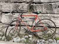 Full side profile of the orange Peugeot road bike showing frame, tires, and saddle in outdoor setting on rocky ground.