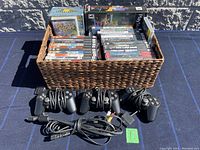 Overview of basket containing various PlayStation 2 game cases and boxes, alongside four PlayStation 2 controllers and connecting cables on a blue surface.