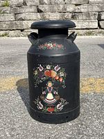 Handpainted black metal milk can standing upright on pavement, showing detailed folk art of a woman, hearts, flowers and birds on the body.