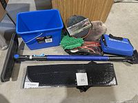 Blue plastic bucket, ice scraper with long blue handle, two cleaning mitts (green chenille microfiber, brown plush), emergency booster cables in clear zippered bag, blue soft case labeled 'Premiers Soins', black sequined case, black telescoping handle part.