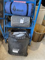 Blue shelving unit with three camping item bags: Woods sleeping bag, JJ Cole ground cover, and Coleman queen air mattress storage bag